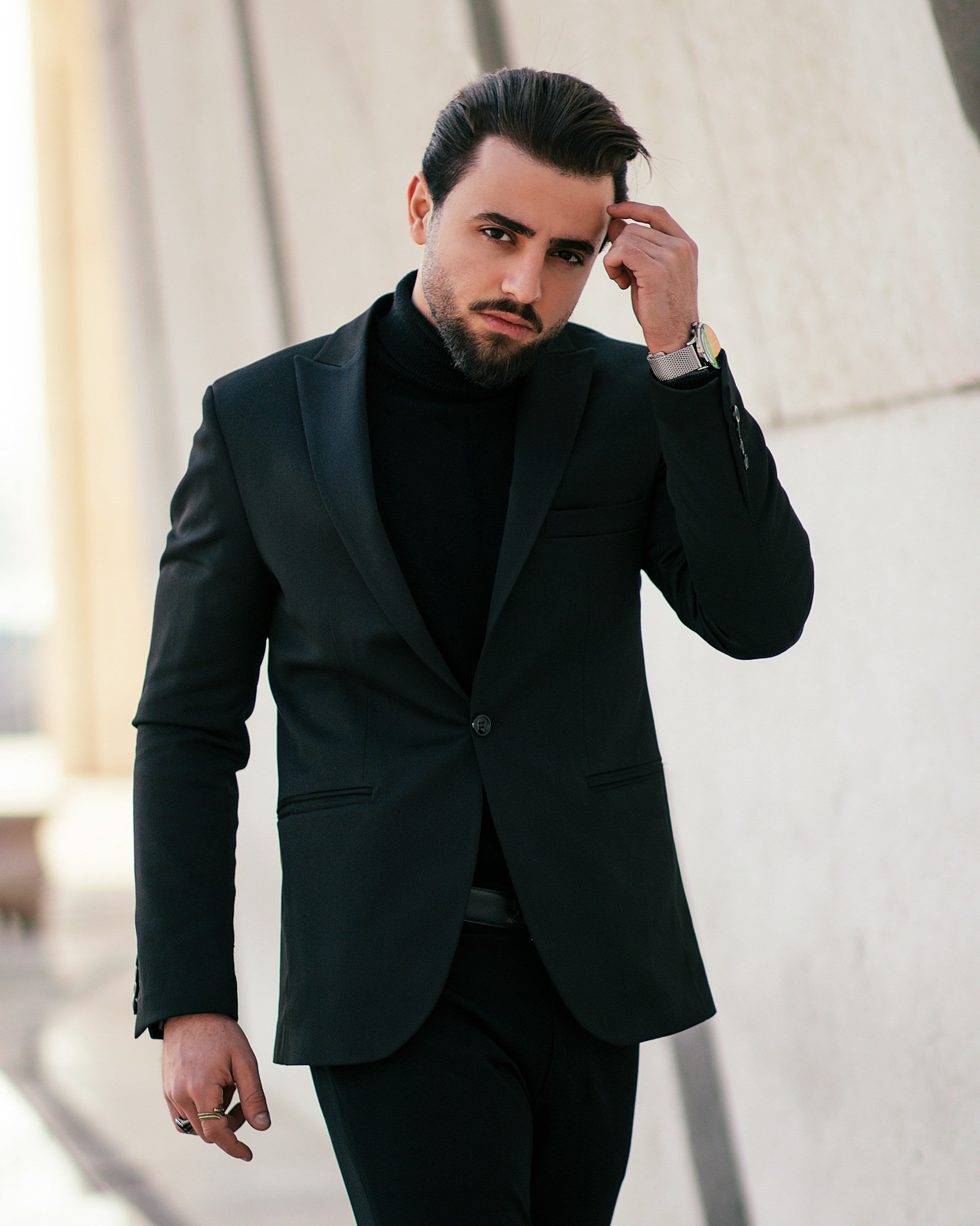 Stylish man in a black suit and turtleneck walks confidently, adjusting his hair against a modern architectural backdrop.