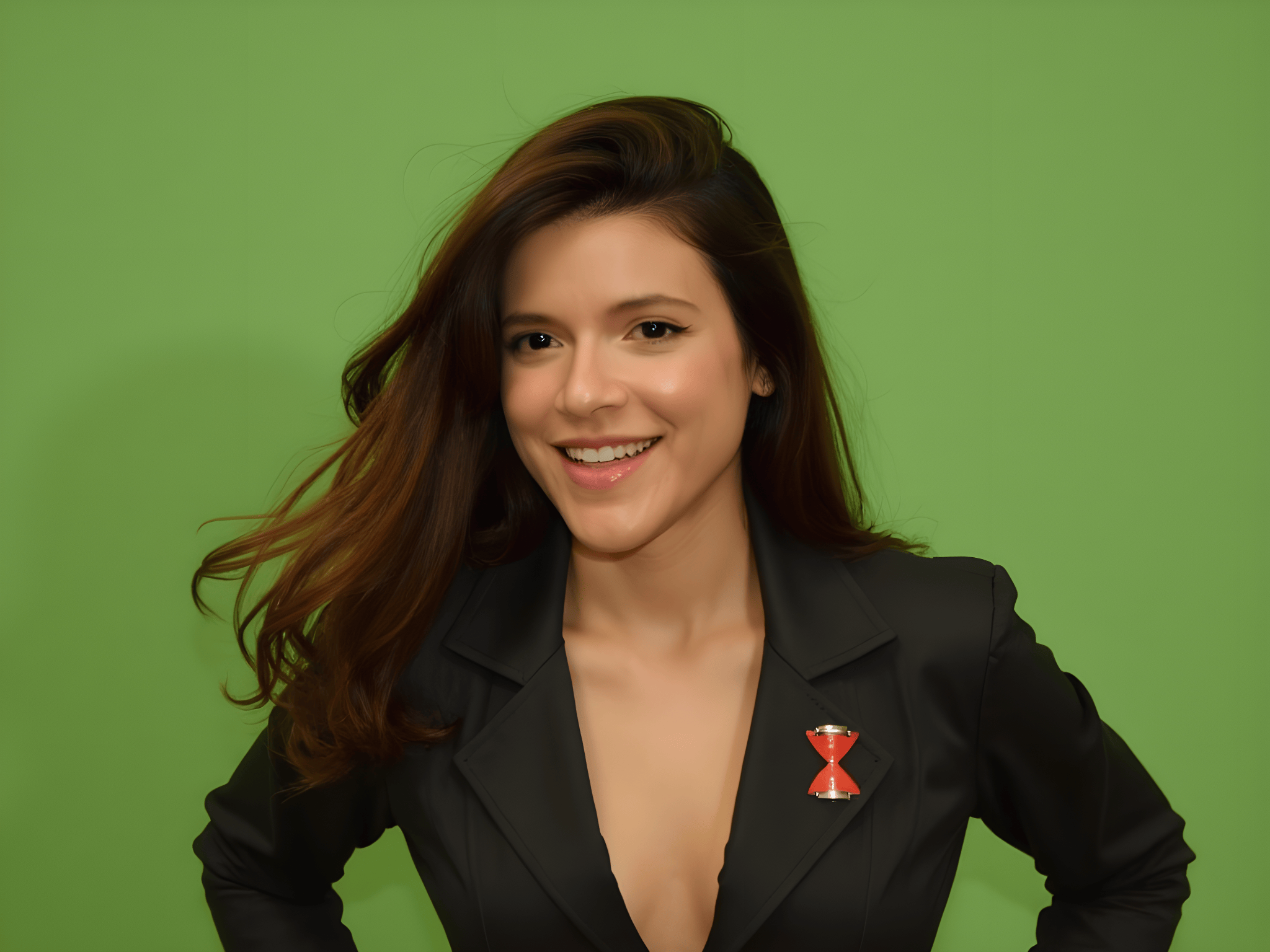 Smiling woman with long hair wearing a black blazer and a red hourglass pin against a vibrant green background.