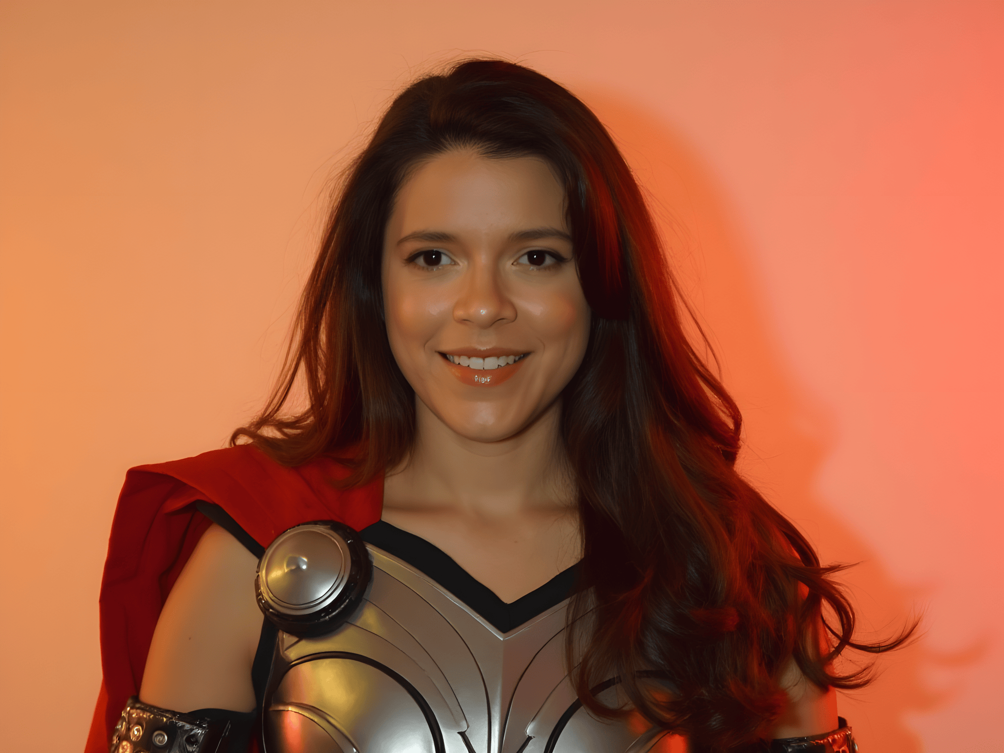 Person dressed in a superhero costume with metallic details and a red cape, smiling against a warm orange background.