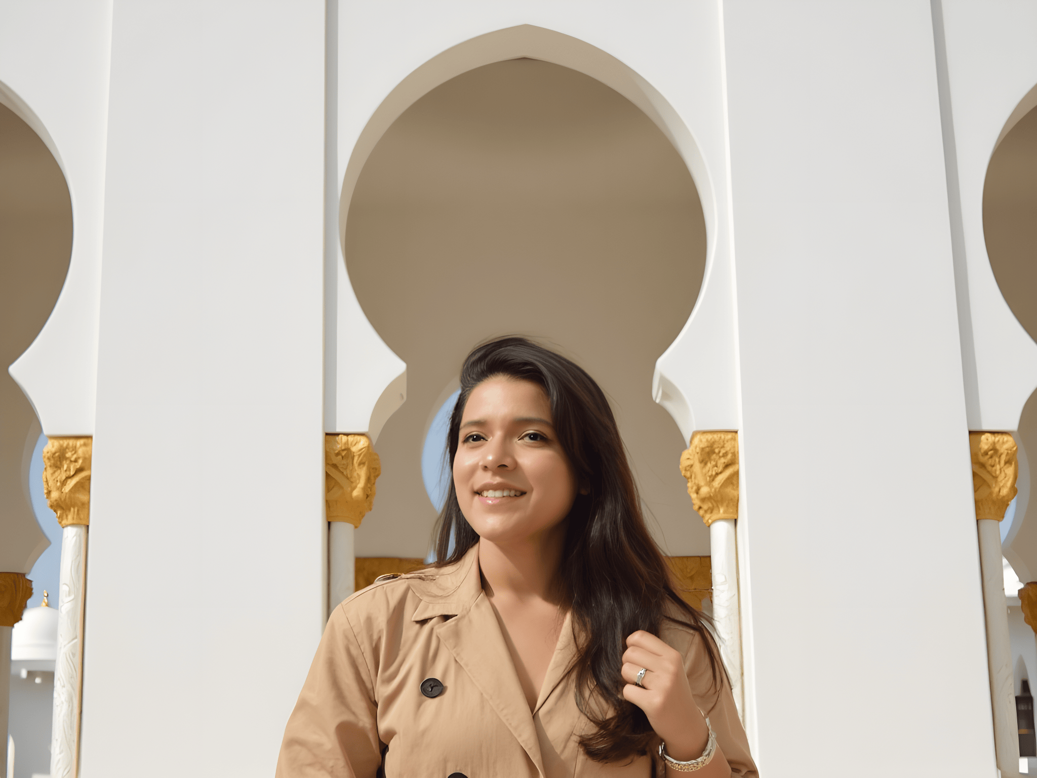 Smiling woman in a beige trench coat posing under ornate arches with golden accents, showcasing a sunny day.