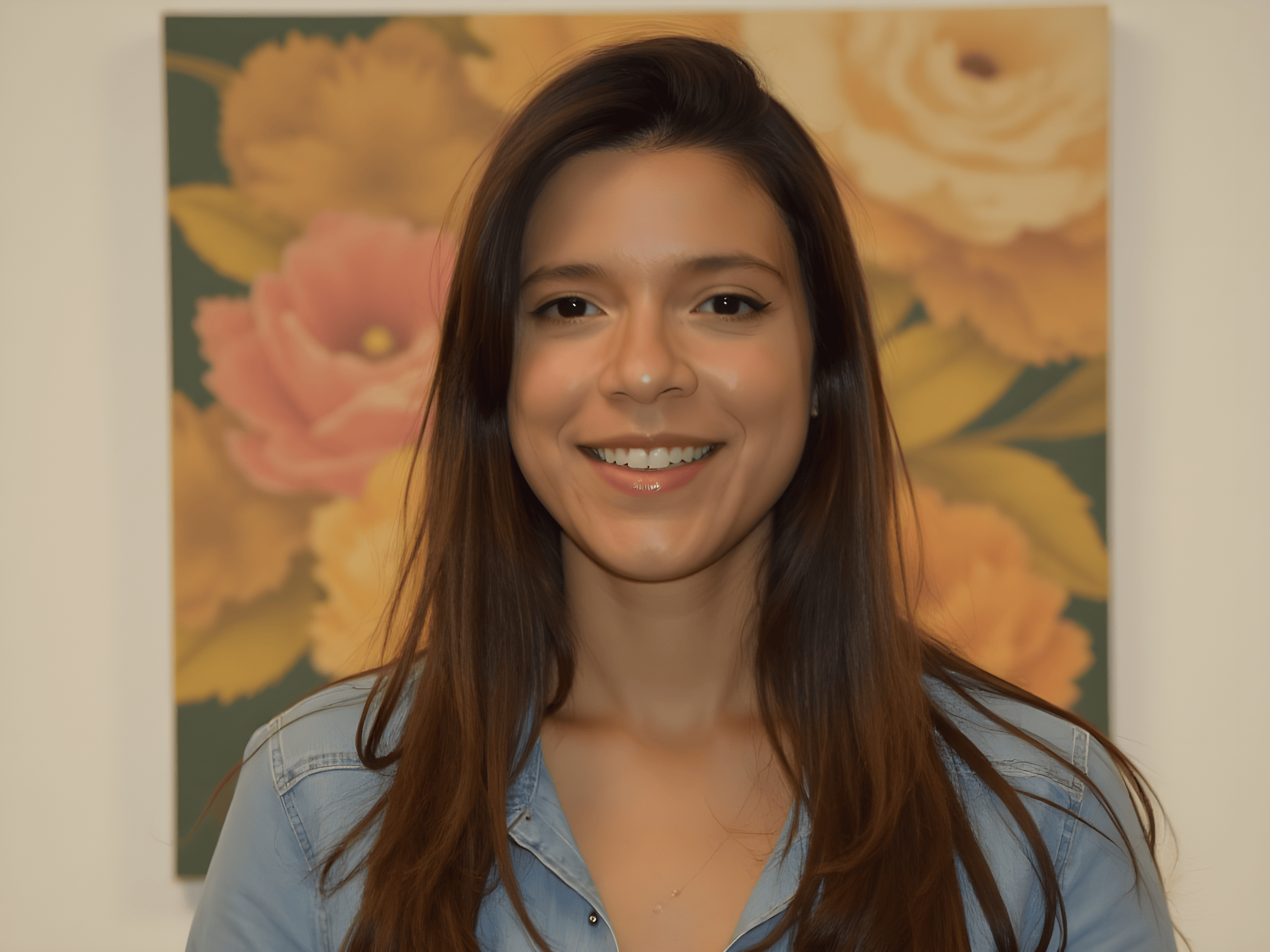 Smiling young woman with long hair wearing a denim shirt, standing in front of floral artwork.
