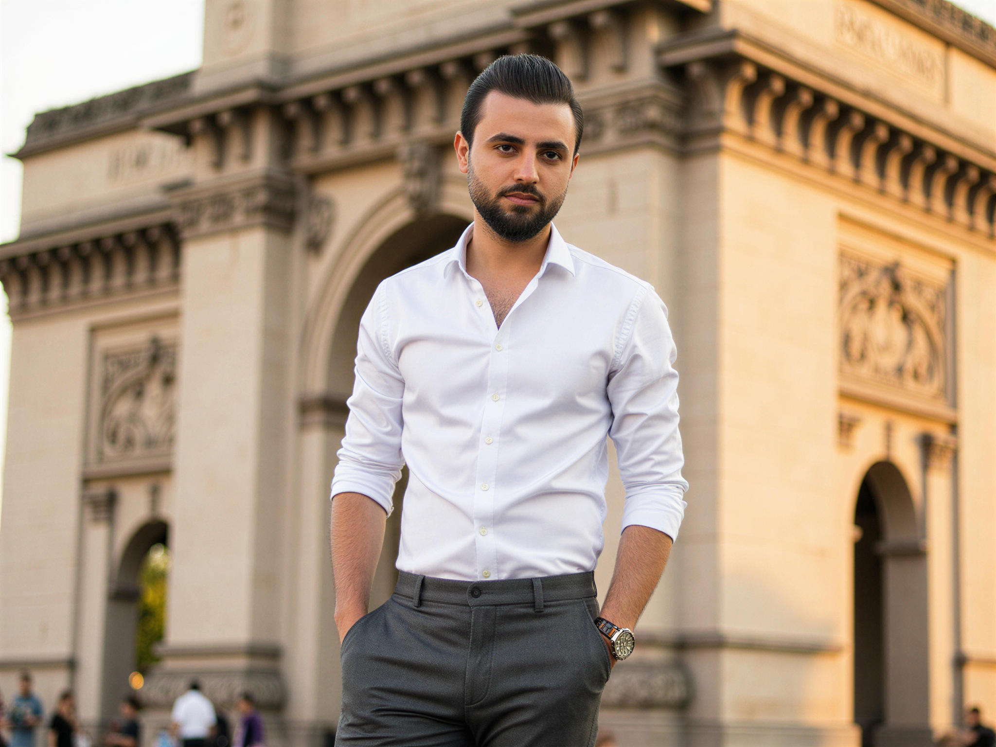 A joyful male figure, OD62S, aged 32, standing in front of the iconic Gateway of India in Mumbai. He wears a tailored smart-casual outfit featuring a crisp white shirt and charcoal grey trousers, exuding effortless sophistication. The light captures the intricate architectural details of the monument behind him, highlighting its majestic arches and historical significance. He has a warm smile that radiates happiness, while the soft afternoon sunlight casts gentle shadows and enhances the vibrant atmosphere. The composition balances his figure with the grandeur of the Gateway, creating a harmonious blend of culture and modernity, evoking a sense of pride and joy associated with this historic landmark.