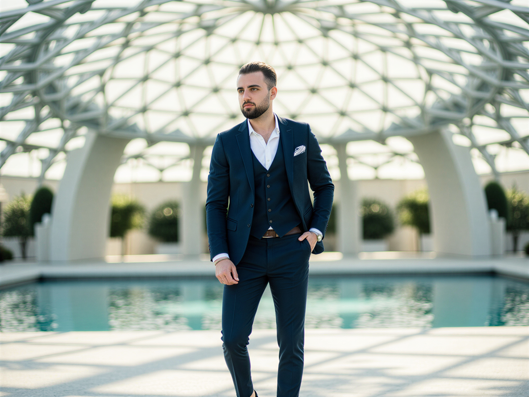 A charismatic male figure, OD62S, aged 32, exuding joy and confidence as he stands in front of the stunning architectural marvel of the Louvre Abu Dhabi. Dressed in a tailored navy suit with a crisp white shirt and minimalist black dress shoes, he gazes up in admiration at the dome's intricate geometric patterns. The sunlight filters through the dome, casting beautiful shadows around him, enhancing the vibrant atmosphere. His relaxed yet sophisticated posture, with one hand casually tucked in his pocket, reflects a happy and open demeanor, embodying the spirit of contemporary cultural appreciation. The backdrop features the museum's serene water mirror, symbolizing clarity and artistic reflection.