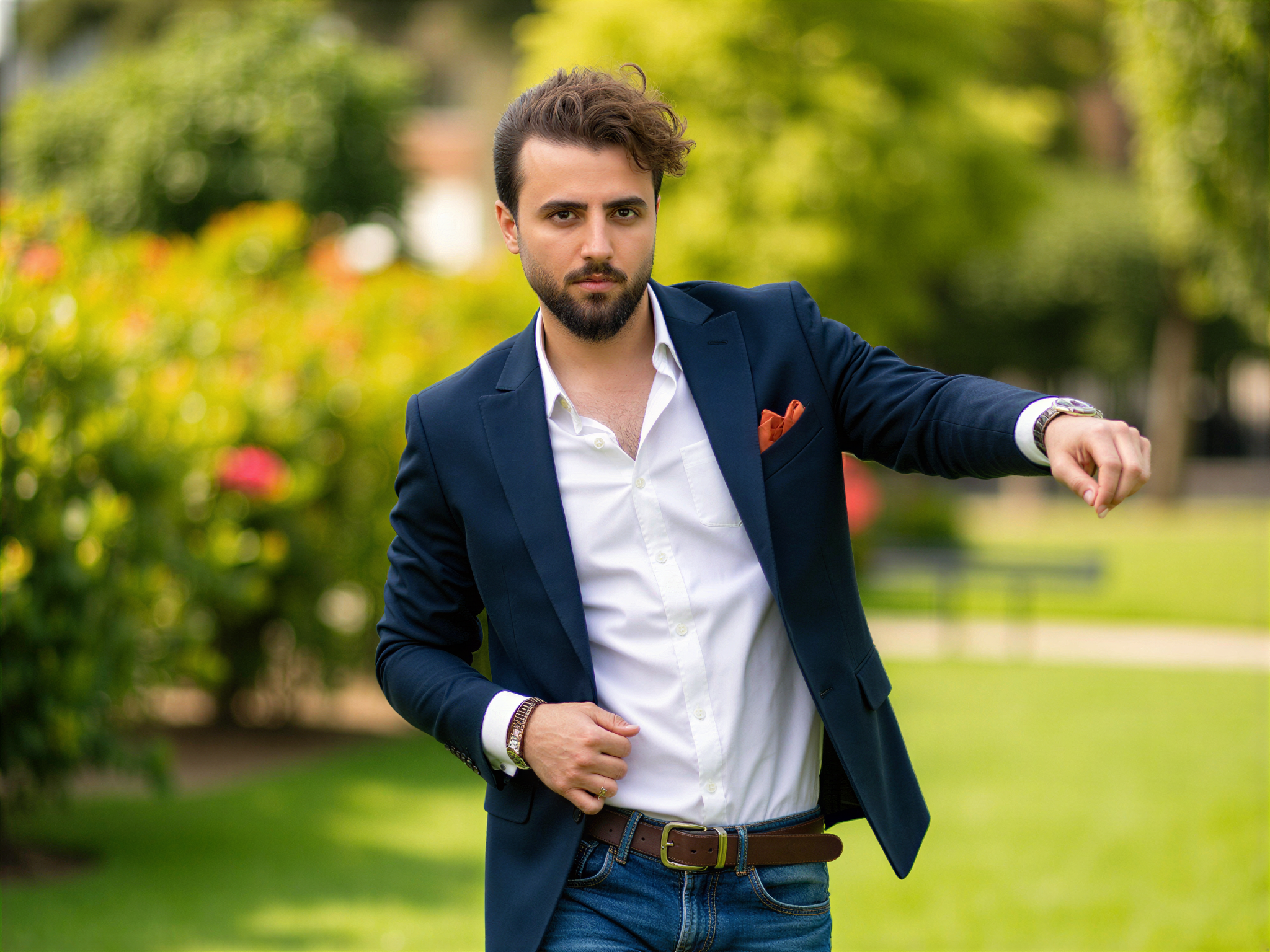 A 32-year-old male, OD62S, captured in a lively, outdoor portrait in the UK. He stands jubilantly in a sun-drenched park, wearing a tailored navy blazer over a crisp white shirt, with the top button playfully undone. His denim jeans add a casual vibe, while a light breeze tousles his hair, enhancing the sense of joy and freedom. The background features lush greenery and blooming flowers, creating a vibrant, cheerful atmosphere. His genuine smile radiates happiness, inviting viewers into the moment, perfectly embodying a spontaneous and carefree spirit. The composition utilizes natural lighting to illuminate his features, highlighting the warmth of his expression.