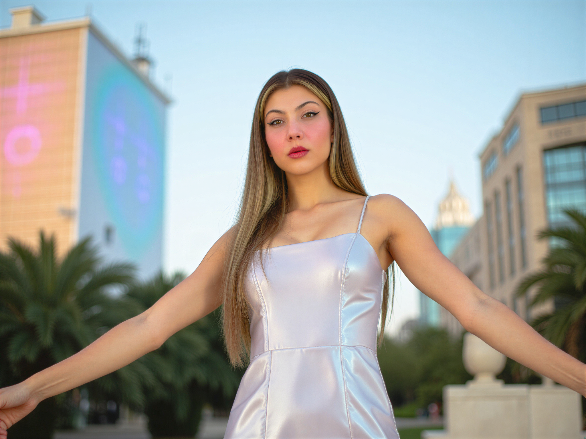 A futuristic portrait of a female individual, OD62S, aged 28, embodying a sense of joy and optimism about the future. She stands center-stage in a sleek, iridescent gown that shifts colors with the light, reflecting hues of silver and lavender. Her makeup is bold yet radiant, featuring metallic accents and glossy lips. Surrounding her are elements of a utopian cityscape, with luminescent buildings and vibrant greenery intermingling, bathed in soft, pastel hues. The lighting is bright and airy, creating an uplifting ambiance. Her pose is dynamic, arms outstretched in a gesture of celebration, radiating happiness and hope for what lies ahead. This visual narrative encapsulates a vision of a harmonious and vibrant future.