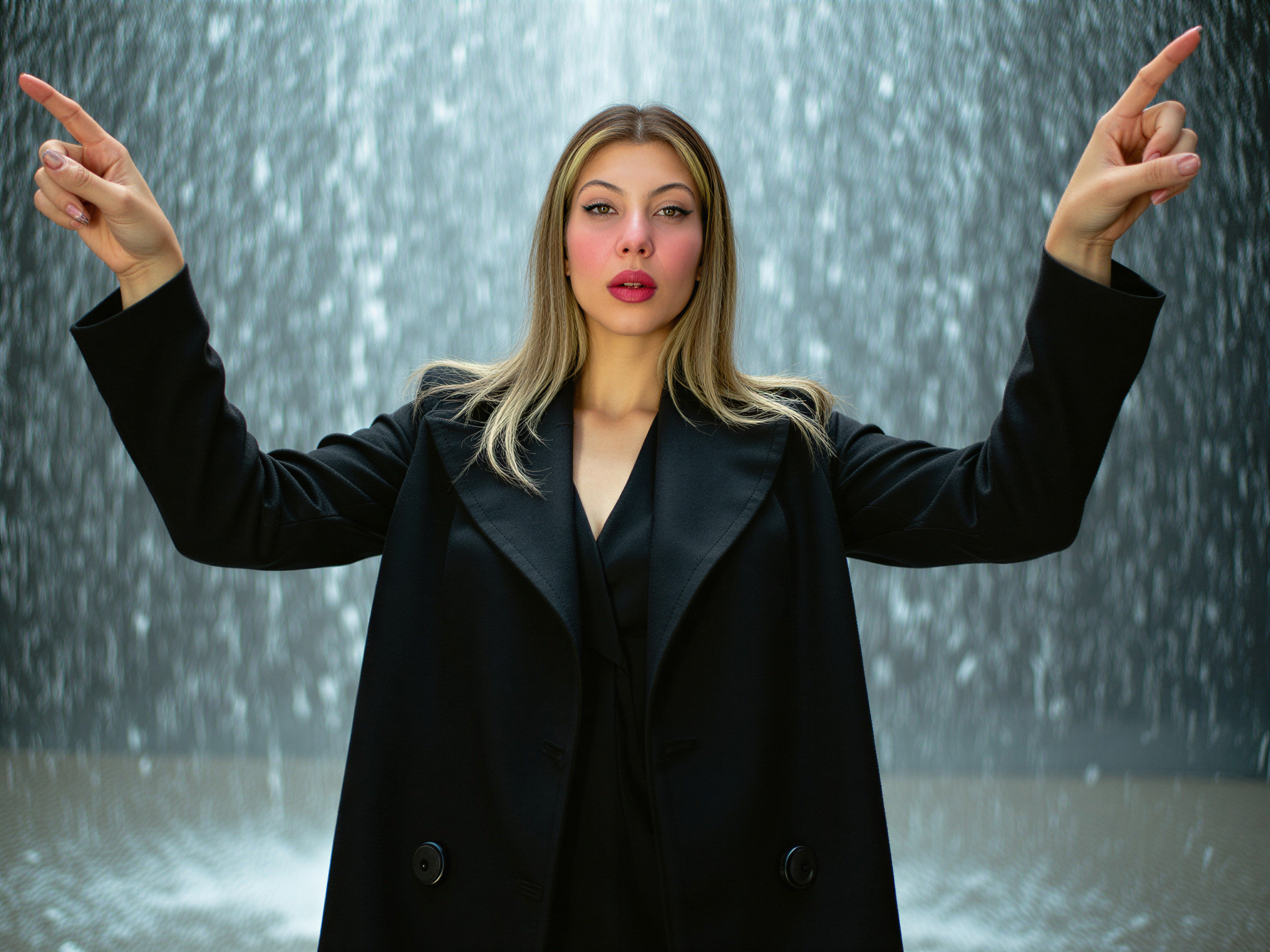 A female subject, OD62S, aged 28, is joyfully experiencing a Rain Room installation, where cascading water creates ethereal reflections and a mesmerizing atmosphere. She is dressed in a chic, fitted black trench coat that contrasts beautifully with the shimmering water surrounding her. Her hair, slightly damp yet elegantly tousled, frames her face as she gleefully lifts her arms, embracing the rain with a beaming smile. The soft, diffused lighting captures the droplets glistening like diamonds, enhancing the serene yet vibrant mood of happiness. The setting evokes a sense of playful liberation, capturing a moment of pure joy in an artistic, immersive environment.