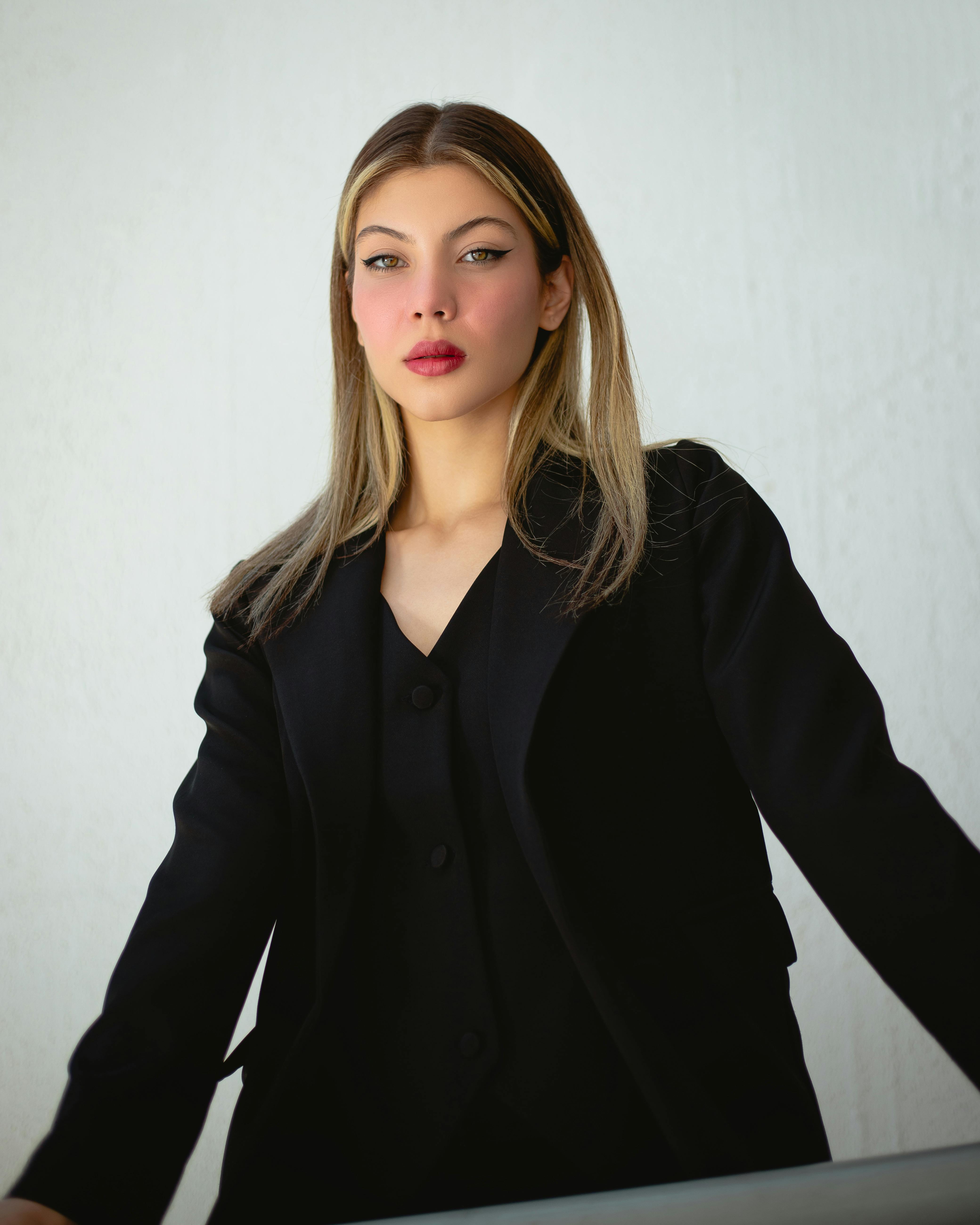 Young woman with stylish long hair and bold makeup, wearing a chic black suit, posing against a minimalist white background.