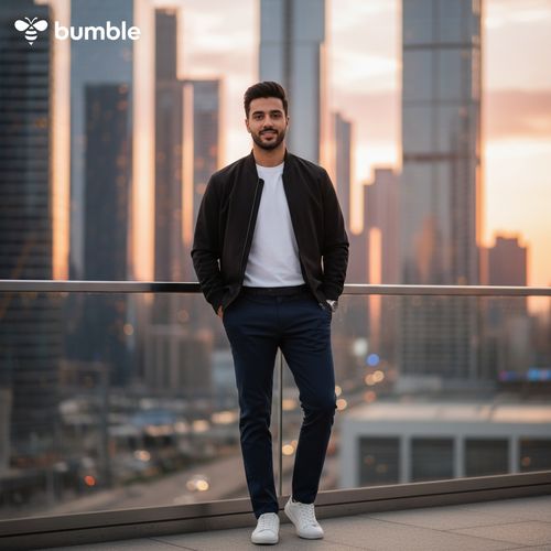 A striking 29-year-old Arab male, David, effortlessly embodies the spirit of Bumble in an urban setting. He is styled in a casual yet sophisticated outfit: a tailored black bomber jacket layered over a fitted white tee, complemented by slim-fit navy chinos. His confident stance exudes approachability, with a subtle smile that invites connection. The backdrop is a vibrant cityscape, with soft pastel hues of sunset reflecting off glass buildings, symbolizing opportunity and warmth. The shot captures a blend of dynamic energy and modern romance, emphasizing David's charismatic presence against this lively environment, all under natural golden hour lighting. This image radiates the essence of connection and confidence, echoing the Bumble ethos.