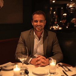 David, a 40-year-old male, is sitting at a cozy, upscale restaurant table set for a romantic dinner. He is casually dressed in a stylish shirt and blazer, smiling warmly towards the camera. The setting is intimate with soft ambient lighting, candles on the table, and a subtle backdrop of elegant décor, creating a perfect atmosphere for dating.
