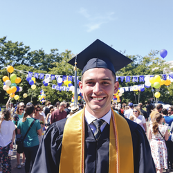 David, a 25-year-old male graduate, smiling brightly at his graduation ceremony. He is wearing a traditional cap and gown, standing outdoors with friends and family in the background enjoying the celebration. The setting is filled with balloons and banners, capturing a joyful and momentous occasion, with a clear blue sky above.
