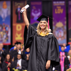 Olivia, a 30-year-old female, dressed in an elegant graduation gown and cap, standing proudly on stage at her graduation ceremony. The setting is vibrant, filled with happy graduates and applauding family members. Olivia is holding her diploma aloft, showcasing her achievement, with a backdrop of colorful banners celebrating the graduation event.