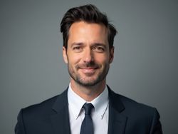 Close-up portrait of a confident businessman in a dark suit and tie, smiling against a neutral gray background.