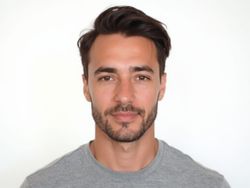 Portrait of a man with short dark hair and a light gray shirt, smiling against a plain white background.