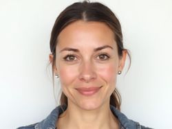Smiling woman with medium brown hair wearing a denim jacket and simple earrings, against a neutral background.