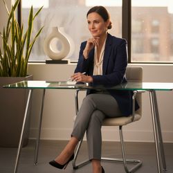 Professional businesswoman seated at a glass desk in a modern office, wearing a navy blazer and light blouse, with green plants and a decorative vase in the background.