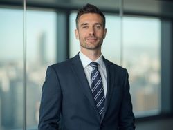 James, a professional male in his 30s, is dressed in a tailored suit with a crisp white shirt and a stylish tie, standing confidently in a modern office setting. The background features a sleek glass window with a cityscape visible outside, hinting at an urban environment. The lighting is bright and professional, enhancing the sophisticated atmosphere of the office.