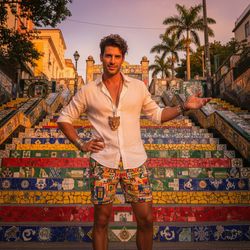 Michael, a 33-year-old male model, striking a confident pose atop the colorful mosaic staircase of Rio de Janeiro's Selarón Steps during golden hour. He wears a tailored white linen shirt that contrasts with his sun-kissed skin and vibrant patterned shorts that encapsulate the spirit of Carnaval. His tousled hair and playful smirk highlight his charisma, while artisan jewelry adds a cultural flair. The warm light accentuates the vibrant colors of the tiles and his attire, creating an atmosphere of joy and freedom, celebrating the excitement of Rio's Carnaval.