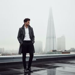 David, a 29-year-old male model, striking a pose on a misty London rooftop, showcases contemporary British style. He is dressed in a fitted charcoal-grey wool trench coat with tailored lapels over a crisp white dress shirt and slim black trousers, embodying sophistication. His polished black leather Oxfords complete the look. With tousled dark hair and a sharp jawline, he channels a modern James Dean vibe. The atmospheric backdrop features the iconic Shard piercing through soft fog. The subdued lighting enhances the melancholic beauty of the scene, as David gazes into the distance with a contemplative expression, embodying urban exploration and refined sophistication. The composition follows the rule of thirds, creating a visually engaging dynamic that reflects the emotional complexity of the modern British man. The editorial image speaks to style, identity, and the contemporary cultural landscape in the UK.
