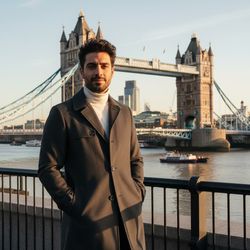 Michael, a 28-year-old Middle Eastern male, stands confidently before the iconic Tower Bridge in London, dressed in a sharply tailored charcoal grey trench coat over a fitted white turtleneck. The golden hour glow creates a cinematic ambiance, highlighting his rugged jawline and tousled hair. His pose is relaxed yet purposeful, with one hand in his pocket, reflecting urban sophistication. The majestic Tower Bridge frames the scene beautifully, blending classic and modern elements, evoking a sense of adventure and ambition in contemporary London life. This image captures not just a moment, but a narrative of exploration and style.