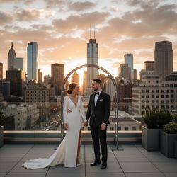 Bride and groom smiling and holding hands on a rooftop during sunset, with a city skyline in the background featuring tall buildings and a cloudy sky.