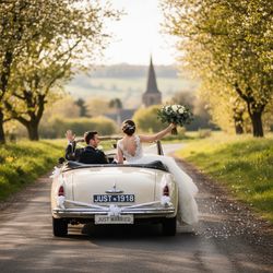 A newlywed couple joyfully rides in a vintage convertible car down a scenic country road, with the bride holding a bouquet and waving while the groom drives, surrounded by lush greenery and a church in the background.
